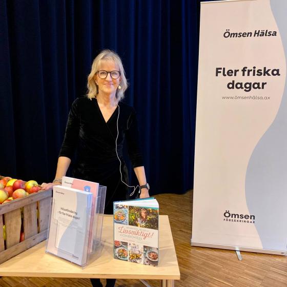 Mai-Lis Hellenius står bakom ett bord vid föreläsningstillfället. På bordet finns broschyrer och äpplen.