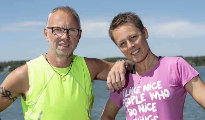 Stefan och Fia står utomhus med vatten i bakgrunden. Stefan lutar sig mot Fia.