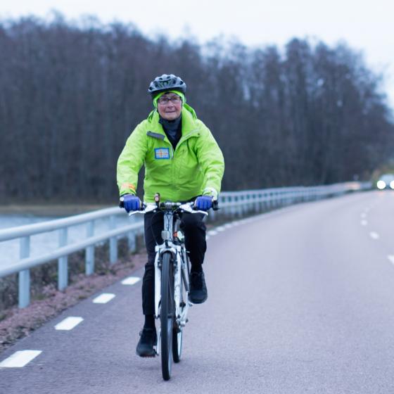 Marita Selander på cykel Marita cyklar vid vägkanten på en trafikerad väg en mulen dag.